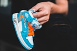 A hand holds a small, vibrant sneaker with a blue and white color scheme and bright orange laces. The sneaker features branding elements and detailed stitching.
