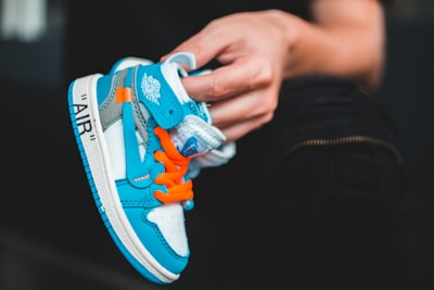 A hand holds a small, vibrant sneaker with a blue and white color scheme and bright orange laces. The sneaker features branding elements and detailed stitching.