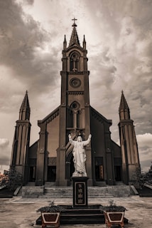 A Gothic-style church with tall, ornate spires and intricate architectural details. In front of the church is a statue of a religious figure with outstretched arms, set against a cloudy, dramatic sky.