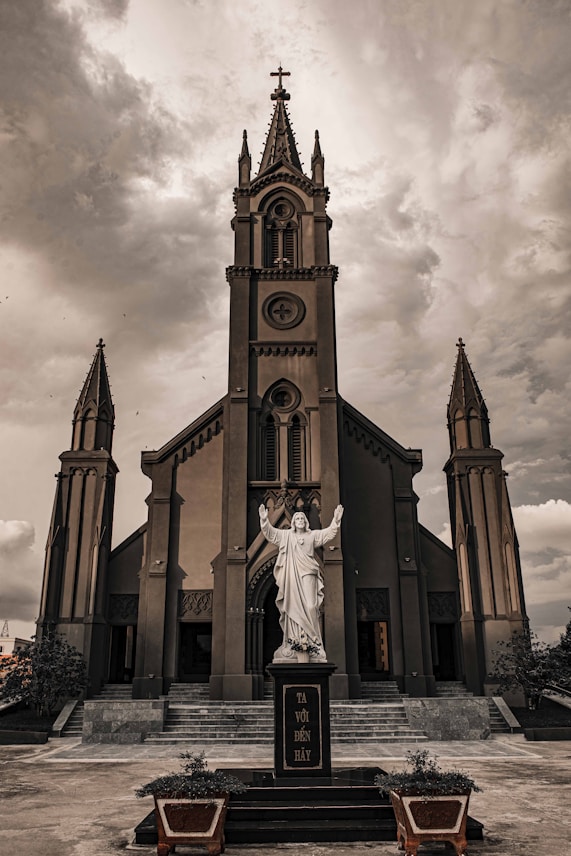 A Gothic-style church with tall, ornate spires and intricate architectural details. In front of the church is a statue of a religious figure with outstretched arms, set against a cloudy, dramatic sky.