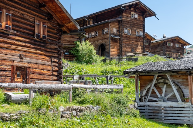 Several rustic wooden houses are set against a lush green hillside on a sunny day. The buildings feature traditional wooden logs and are elevated with small stone walls. A wooden structure on the right, possibly a shed or barn, adds to the rural atmosphere.