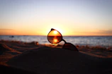 Close-up of a sleek pair of Tapped Sunset sunglasses resting on a sandy beach at dusk.