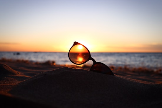 Sunglasses rest on a mound of sand, with the setting sun visible through one lens, casting a warm glow over the beach and ocean horizon.