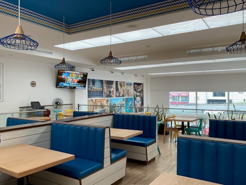 A modern restaurant interior with blue and white seating booths, ceiling light fixtures, and a wooden floor. A television mounted on the wall displays news, while the walls feature decorative photos. Large windows let in natural light, and there are plants along the railing.