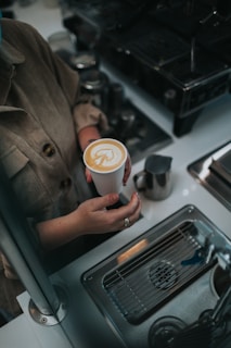 A person holds a cup of coffee with intricate latte art on the surface. The scene is set in a café environment with a metal sink, espresso machine, and various metal containers visible.