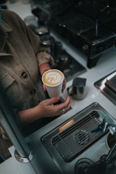 A person holds a cup of coffee with intricate latte art on the surface. The scene is set in a café environment with a metal sink, espresso machine, and various metal containers visible.