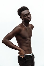 A fit young man poses shirtless against a plain white background, displaying a confident posture with one hand resting on his hip. He has short, curly hair and a neutral expression on his face.