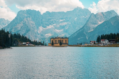 A serene alpine lake with clear blue waters is surrounded by tall, majestic mountains. A distinctive, large hotel building with a yellow facade is located near the edge of the lake, and smaller structures are visible nearby, nestled among evergreen trees. The sky is partly cloudy, casting a soft, diffused light over the landscape.