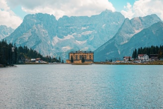 A serene alpine lake with clear blue waters is surrounded by tall, majestic mountains. A distinctive, large hotel building with a yellow facade is located near the edge of the lake, and smaller structures are visible nearby, nestled among evergreen trees. The sky is partly cloudy, casting a soft, diffused light over the landscape.