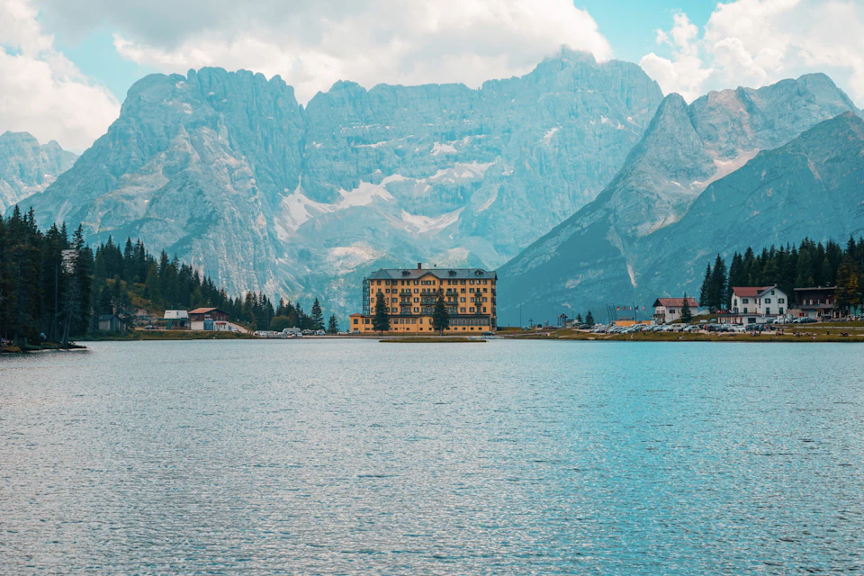 The image depicts a serene lake surrounded by majestic mountains, with a large yellow building situated on the shore. The calm waters of the lake reflect the clear blue sky above, creating a picturesque scene. In the distance, several small houses can be seen nestled among the mountains, adding to the tranquil atmosphere.
