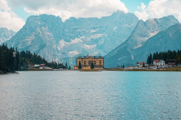 A serene alpine lake with clear blue waters is surrounded by tall, majestic mountains. A distinctive, large hotel building with a yellow facade is located near the edge of the lake, and smaller structures are visible nearby, nestled among evergreen trees. The sky is partly cloudy, casting a soft, diffused light over the landscape.