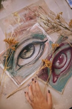 An artistic composition featuring painted sketches of eyes in shades of blue and maroon placed on a textured surface. Sprigs of dried flowers lay scattered around, adding a natural element. A hand with painted nails is positioned in the lower part of the image, gently touching one of the sketches.