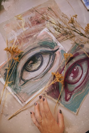 An artistic composition featuring painted sketches of eyes in shades of blue and maroon placed on a textured surface. Sprigs of dried flowers lay scattered around, adding a natural element. A hand with painted nails is positioned in the lower part of the image, gently touching one of the sketches.