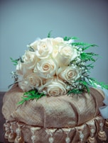 A bouquet of cream-colored roses is elegantly arranged with green foliage, placed atop a decorative base covered in a shimmering gold fabric adorned with tassels and pearls.