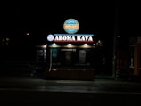 A coffee kiosk with illuminated signage displaying 'Aroma Kava' in large letters. The kiosk is set against a dark background, and its lights cast a bright glow around the structure. Two beverage icons with prices are visible, suggesting available coffee options.