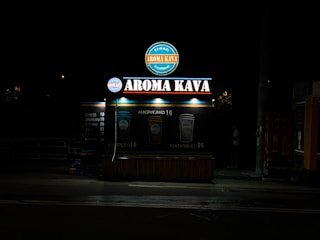 A coffee kiosk with illuminated signage displaying 'Aroma Kava' in large letters. The kiosk is set against a dark background, and its lights cast a bright glow around the structure. Two beverage icons with prices are visible, suggesting available coffee options.