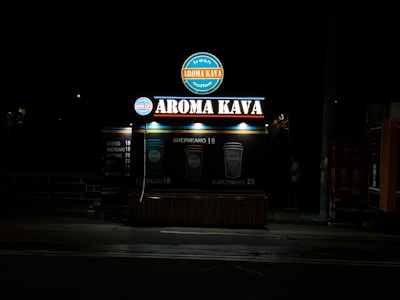 A coffee kiosk with illuminated signage displaying 'Aroma Kava' in large letters. The kiosk is set against a dark background, and its lights cast a bright glow around the structure. Two beverage icons with prices are visible, suggesting available coffee options.