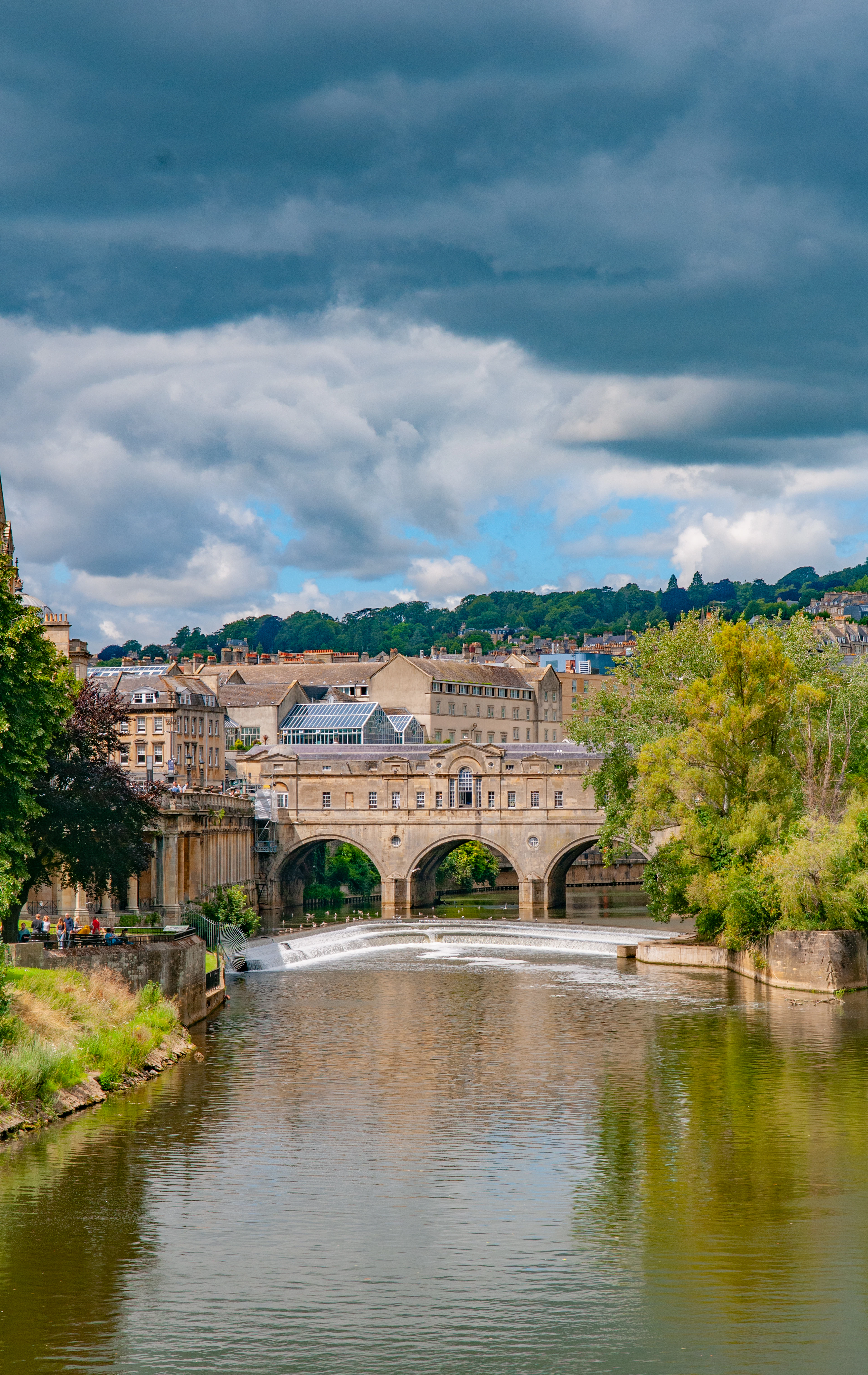 Pulteney Bridge and the horseshoe weir on the River Avon in Bath, framed by trees and honey-stone buildings.