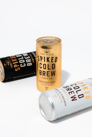 Three cans of Café Agave Spiked Cold Brew coffee are arranged on a white surface. The central can is gold, while the other two are black and white, creating a bright and modern composition.
