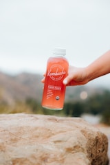 A close-up of a hand pouring kombucha into a glass, with natural ingredients visible in the background.