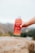 A hand holding a bottle of Kombucha labeled 'Aloha Punch'. The bottle is held over a large rock, with an outdoor, blurry background indicating nature or a scenic view.