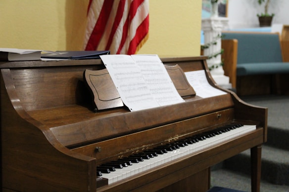 A wooden upright piano with a set of sheet music placed on its music rest. The background shows an American flag, a yellow wall, and some seating that appears to be in a church or a formal gathering space.