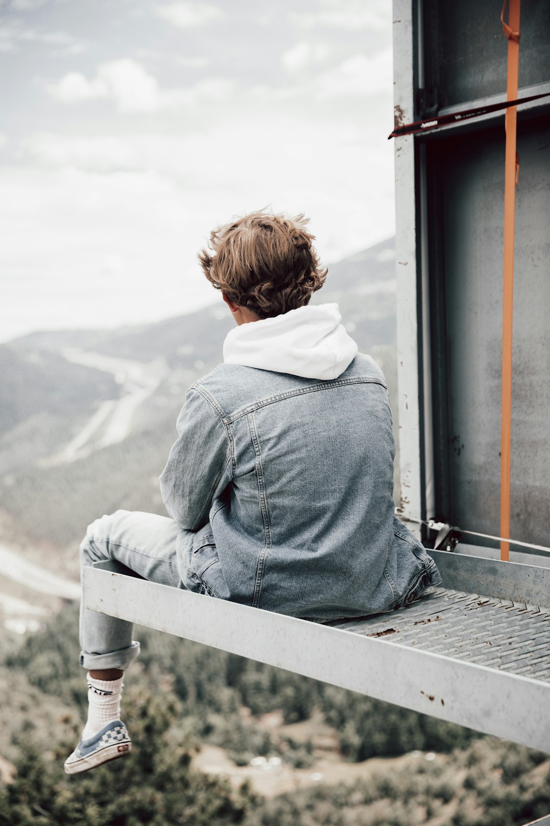 boy in gray hoodie sitting on white wooden fence looking at mountains during daytime