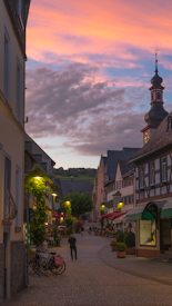 A picturesque European street scene at sunset, featuring cobblestone paths, charming half-timbered buildings, and warm street lighting. A few people stroll leisurely down the street, past outdoor cafe tables adorned with plants. The sky is filled with vibrant pink and orange hues as the sun sets.