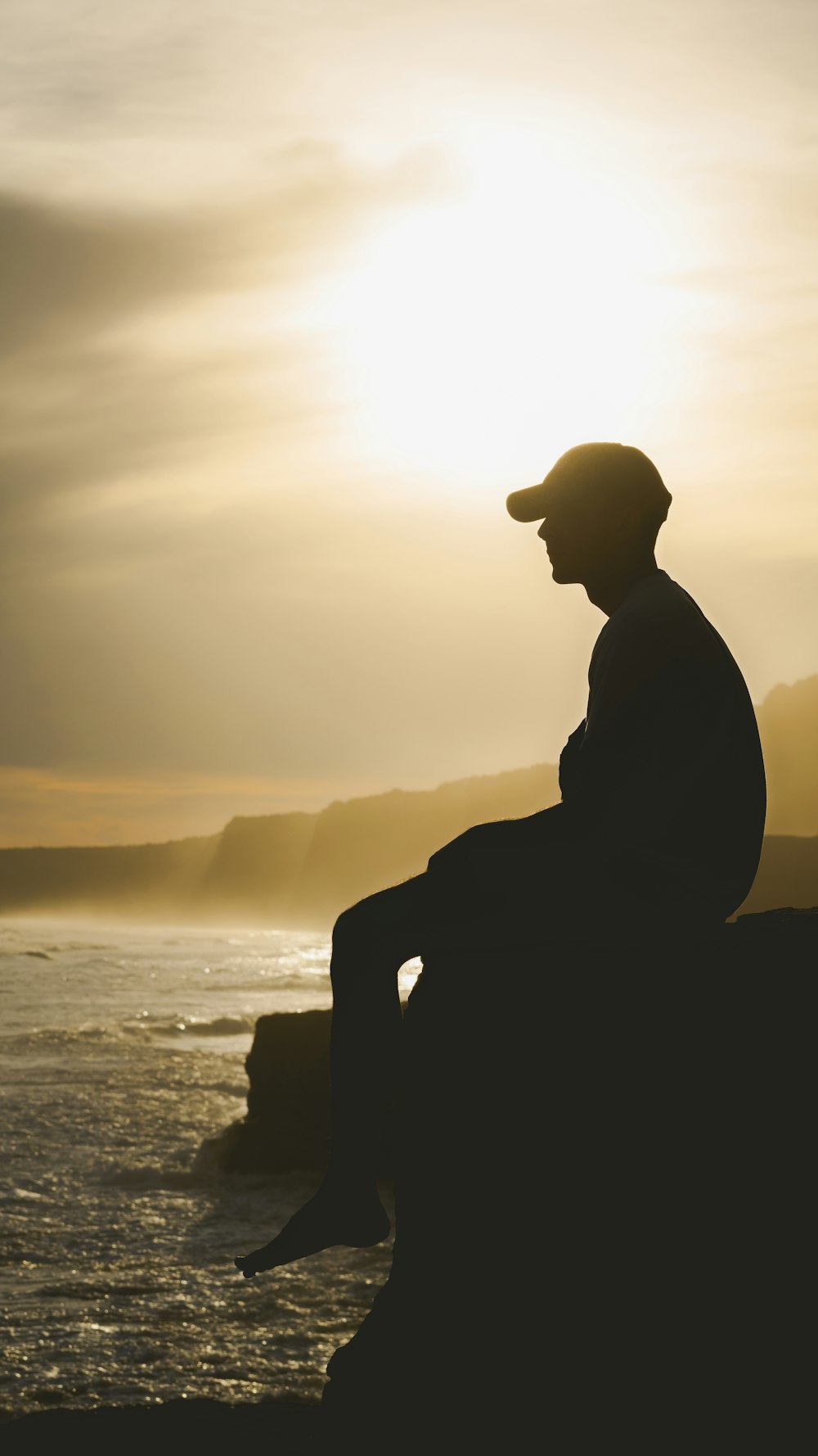 silhouette of man sitting on rock near body of water during sunset