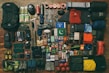 A collection of camping and hiking gear neatly arranged, including backpacks, climbing shoes, sleeping bags, clothing, cooking utensils, safety equipment, and various personal items such as books and snacks. The gear is laid out on a wooden floor, organized by type and color.