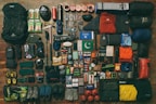 A collection of camping and hiking gear neatly arranged, including backpacks, climbing shoes, sleeping bags, clothing, cooking utensils, safety equipment, and various personal items such as books and snacks. The gear is laid out on a wooden floor, organized by type and color.