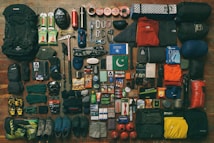 A collection of camping and hiking gear neatly arranged, including backpacks, climbing shoes, sleeping bags, clothing, cooking utensils, safety equipment, and various personal items such as books and snacks. The gear is laid out on a wooden floor, organized by type and color.