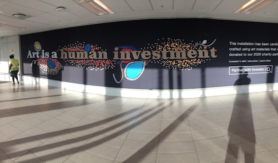 A large mural displayed on a wall inside a building with the words 'Art is a human investment' in bold, artistic lettering. The mural includes colorful abstract designs such as circles and lines surrounding the text. The floor is tiled, and there is a person walking on the left wearing a yellow vest. The installation includes a prominent section of text explaining that it was created using donated art materials.