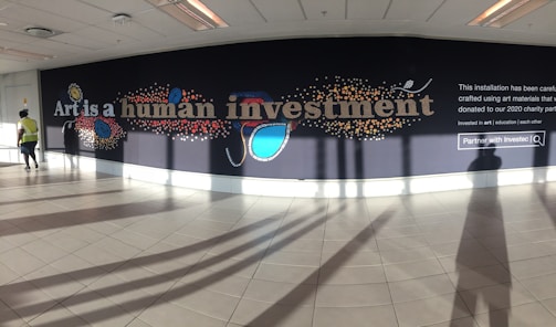 A large mural displayed on a wall inside a building with the words 'Art is a human investment' in bold, artistic lettering. The mural includes colorful abstract designs such as circles and lines surrounding the text. The floor is tiled, and there is a person walking on the left wearing a yellow vest. The installation includes a prominent section of text explaining that it was created using donated art materials.
