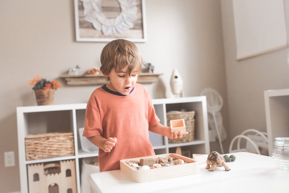 Junge im orangefarbenen Rundhals-T-Shirt vor weißem Holztisch mit Cupcakes