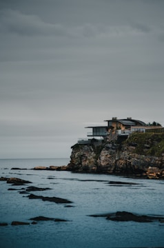 A modern home perched on a cliff overlooking the vast blue Pacific Ocean at sunset.