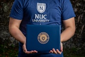 A person is holding a dark blue folder featuring the logo of NUC University. The individual is wearing a matching blue T-shirt with the same university logo and name on the front. The background appears to be natural, possibly with some greenery.