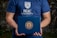 A person is holding a dark blue folder featuring the logo of NUC University. The individual is wearing a matching blue T-shirt with the same university logo and name on the front. The background appears to be natural, possibly with some greenery.