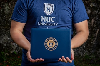 A person is holding a dark blue folder featuring the logo of NUC University. The individual is wearing a matching blue T-shirt with the same university logo and name on the front. The background appears to be natural, possibly with some greenery.