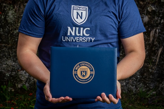 A person is holding a dark blue folder featuring the logo of NUC University. The individual is wearing a matching blue T-shirt with the same university logo and name on the front. The background appears to be natural, possibly with some greenery.