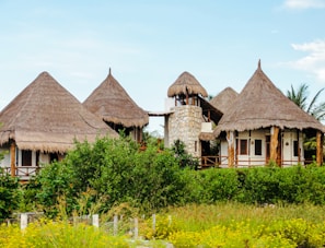 A picturesque setting featuring rustic structures with thatched roofs, surrounded by lush greenery and vibrant yellow flowering plants. The buildings are crafted with natural materials like wood and stone, creating a harmonious blend with nature. The sky overhead is a clear blue, enhancing the tranquil atmosphere.