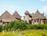 A picturesque setting featuring rustic structures with thatched roofs, surrounded by lush greenery and vibrant yellow flowering plants. The buildings are crafted with natural materials like wood and stone, creating a harmonious blend with nature. The sky overhead is a clear blue, enhancing the tranquil atmosphere.