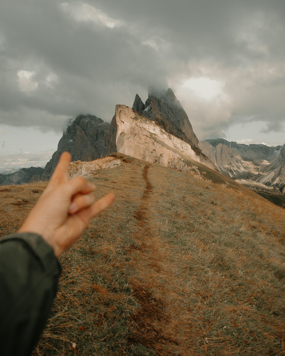 person in black jacket standing on brown grass field near brown rocky mountain under white cloudy