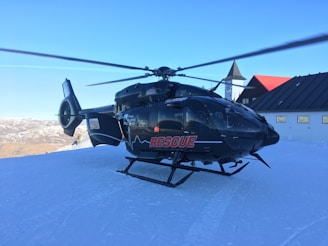 A black rescue helicopter is stationary on a snow-covered surface. The helicopter has 'RESCUE' written in red on the side. The background features snow-capped mountains under a clear blue sky, and there is a building with a pitched roof and a clock tower nearby.