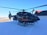 A black rescue helicopter is stationary on a snow-covered surface. The helicopter has 'RESCUE' written in red on the side. The background features snow-capped mountains under a clear blue sky, and there is a building with a pitched roof and a clock tower nearby.