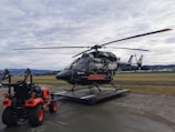 A black helicopter with rescue markings is stationed on a helipad in an outdoor setting. A small orange utility vehicle with visible tires and equipment in the back is positioned next to the helicopter. The sky is overcast, and the surrounding area includes grassy fields and distant hills.