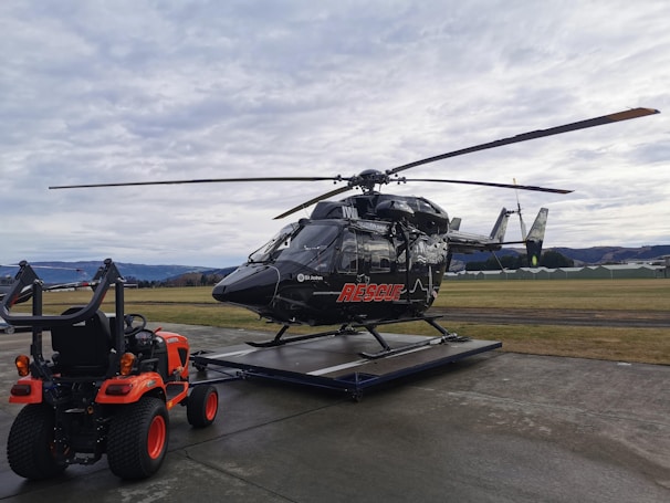 A black helicopter with rescue markings is stationed on a helipad in an outdoor setting. A small orange utility vehicle with visible tires and equipment in the back is positioned next to the helicopter. The sky is overcast, and the surrounding area includes grassy fields and distant hills.