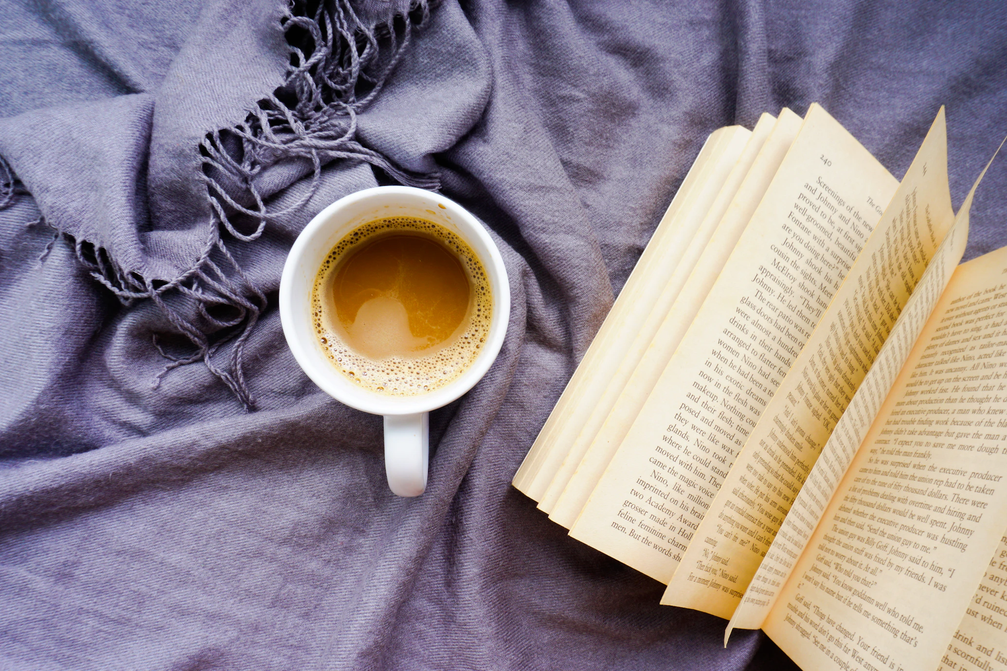Une tasse de café posée sur un tissu violet froissé, à côté d’un livre ouvert aux pages jaunies.