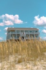 A large, white beach house elevated on stilts sits behind a patch of tall, golden beach grass. The sky above is bright blue with a few fluffy white clouds scattered across.