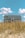 A large, white beach house elevated on stilts sits behind a patch of tall, golden beach grass. The sky above is bright blue with a few fluffy white clouds scattered across.